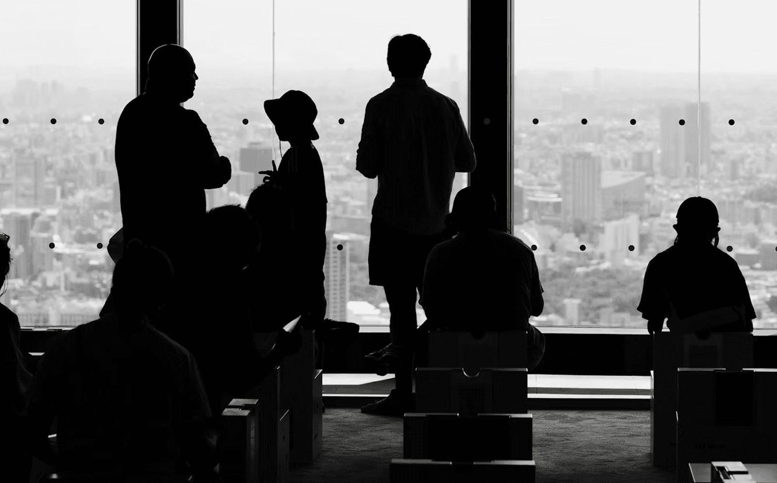 Silhouettes of people standing and sitting near a large window overlooking a cityscape. The scene is in black and white, with the figures in shadow and the city skyline visible through the glass.